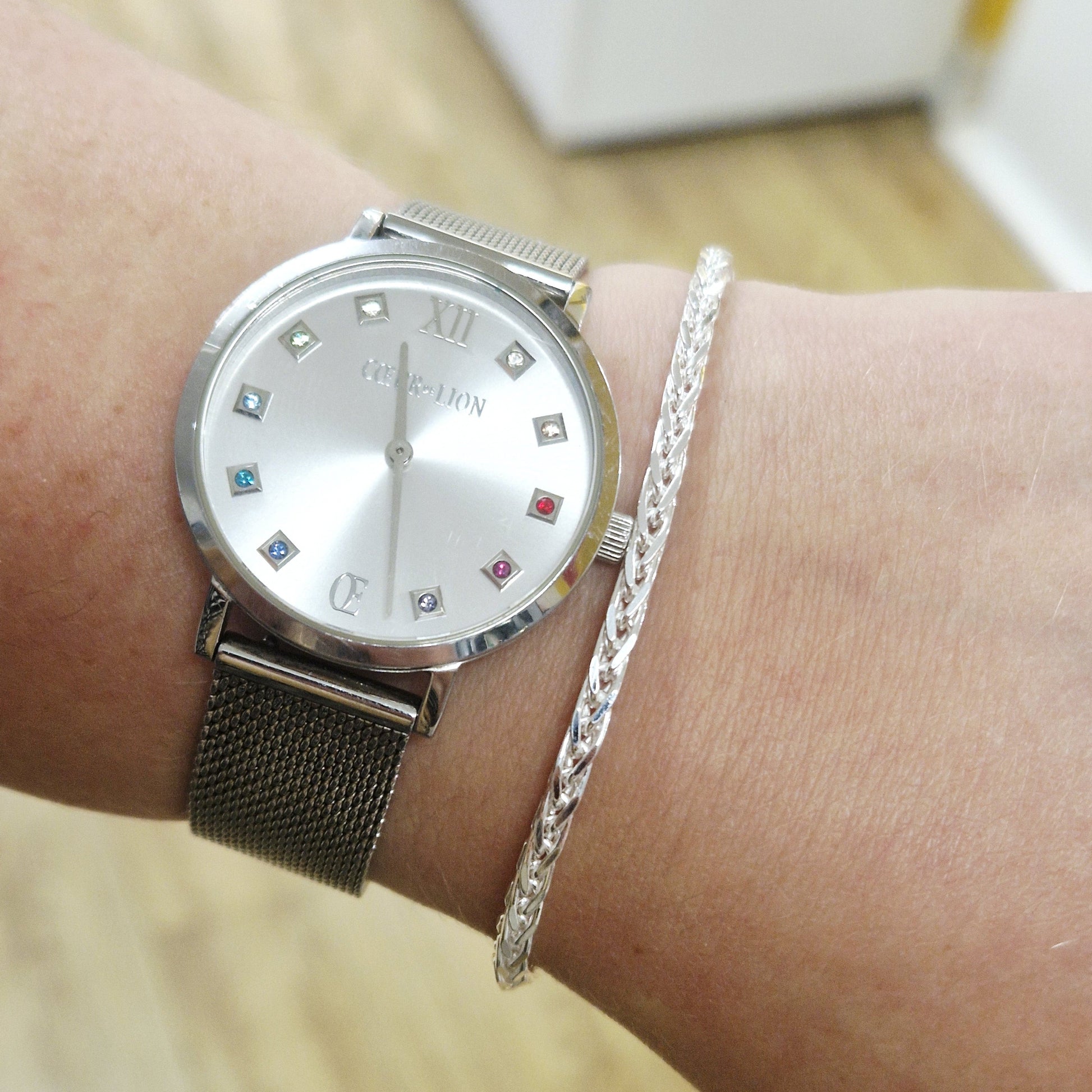 Silver coeur de lion watch with gemstone accents and spiga bracelet on a wrist against a blurred background
