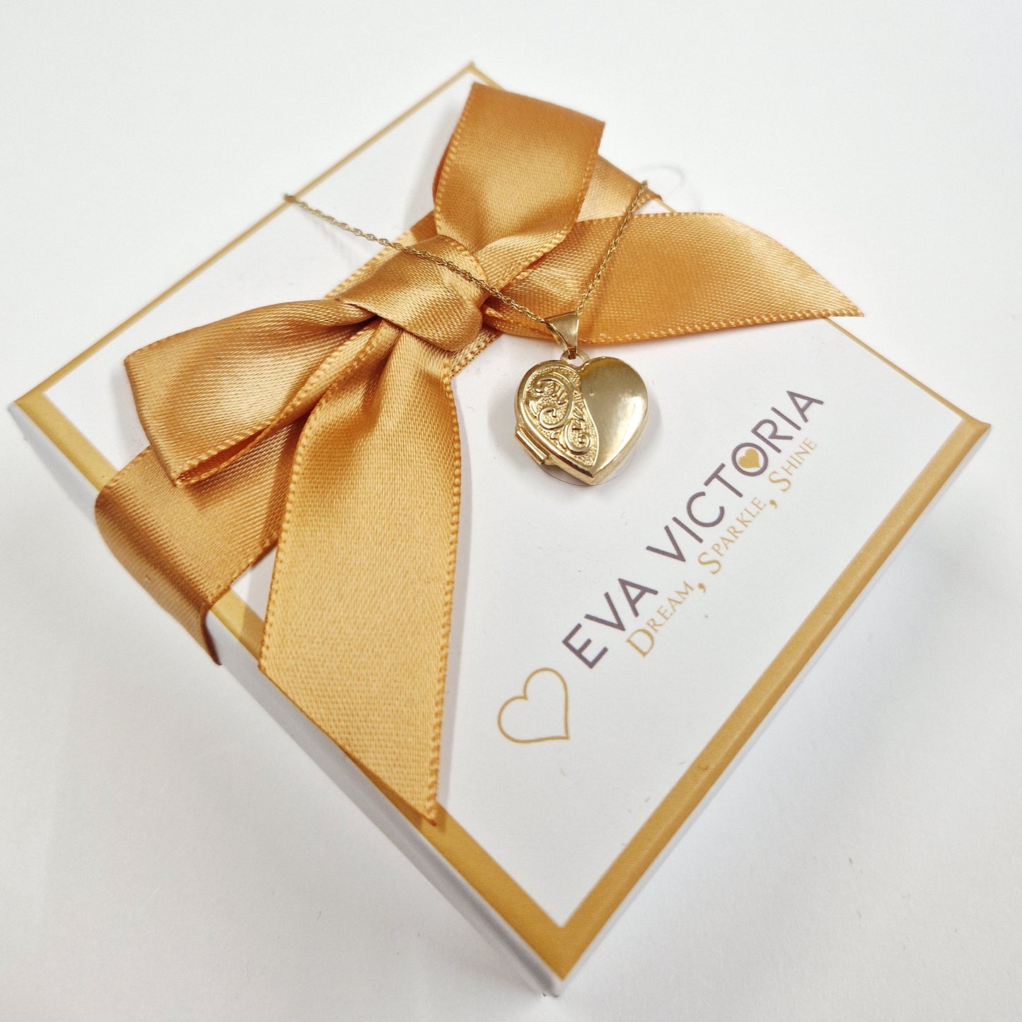 Gold heart-shaped locket on a matching bow with 'EVA VICTORIA' branding on a white background